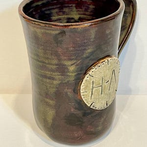 May include: A handmade ceramic mug with a brown, green, and burgundy glaze. The mug features a handle and a circular emblem with the letters "H-A". The mug is approximately 5 inches tall.