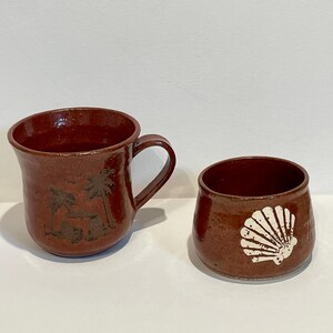 May include: Two brown ceramic mugs. One mug has a handle and a design of palm trees and a hut. The other mug is a small, round cup with a white seashell design. Both mugs have a glossy finish.