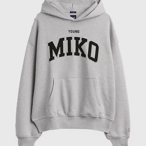 May include: A pale grey hoodie with a drawstring hood and a front pocket. The word "YOUNG" is printed above the word "MIKO" in bold, black letters. The hoodie has long sleeves and ribbed cuffs and hem.