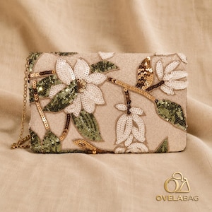 May include: A rectangular clutch bag in a light beige fabric featuring an embroidered floral pattern with white, olive green, and metallic gold sequins. The bag includes a thin gold-toned metal chain strap attached to the side for carrying.