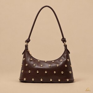 May include: A dark brown shoulder bag featuring a smooth finish and a single strap with knotted ends. The exterior is decorated with evenly spaced gold-tone metal studs across the front. A gold-tone zip closure is visible at the top.