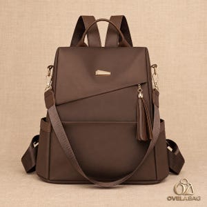 Könnte beinhalten: Ein dunkelbrauner Rucksack mit glatter Oberfläche, einem Tragegriff und verstellbaren Schultergurten. Die Vorderseite zeigt einen goldfarbenen Metallakzent, eine diagonale Reißverschlusstasche und eine braune Quaste. Der Text OVELABAG ist sichtbar.
