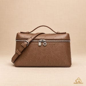 May include: A brown structured vanity bag with a textured ostrich-like pattern, a top handle, and an adjustable shoulder strap. The bag features a silver-toned zipper closure with two metal pulls and the OVELABAG logo printed in the bottom right corner.