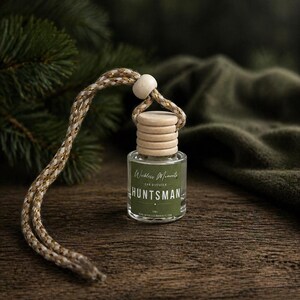 May include: A car air freshener with the text "Huntsman" on a green label. The clear glass bottle has a wooden cap and a braided rope for hanging. The background includes a green fabric and pine branches.