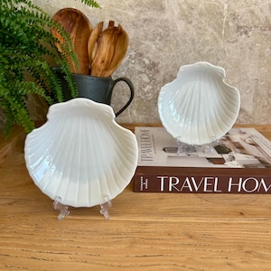 May include: Two white, shell-shaped dishes are displayed on clear stands. The dishes have a glossy finish and a ribbed texture, resembling a scallop shell. The dishes are on a wooden surface, with a book titled "TRAVEL HOME" visible in the background.