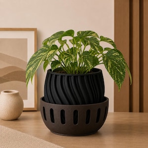 May include: A vibrant green and yellow variegated pothos plant potted in a black, spiraled, cylindrical planter. This planter sits inside a dark brown, bowl-shaped outer pot featuring a series of vertical, oval-shaped cutouts along its base.