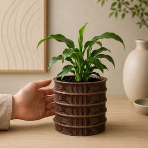May include: A cylindrical brown planter with a ribbed, tiered exterior texture containing a green leafy plant. The planter features a series of horizontal, concave bands that create a repeating pattern around the entire circumference of the vessel.