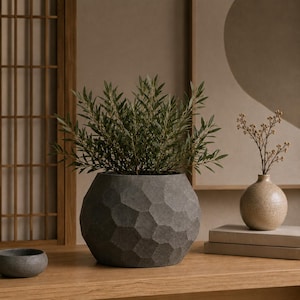 May include: A dark gray, spherical planter with a faceted, geometric honeycomb texture on its exterior surface. The planter holds a lush green plant with thin, pointed leaves. A small, round, beige speckled vase with dried stems sits nearby.