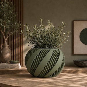 May include: A spherical olive green planter featuring a series of overlapping, diagonal structural bands that create a ribbed, cage-like exterior. The planter holds a lush, leafy green plant with small, pointed foliage extending upwards.