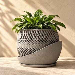 May include: A gray, spherical planter with a textured, lattice-like woven pattern on the upper section and a solid, smooth-curved base. A vibrant green leafy plant is potted inside the container, which sits on a matching circular tray.
