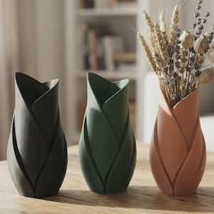 May include: Three vases with a layered petal design, arranged in a row. The first vase is matte black, the second is dark green, and the third is a soft terracotta color, containing a dried floral arrangement of wheat and lavender stems.