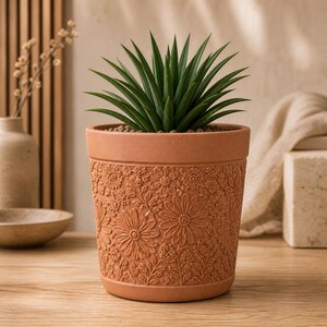 May include: A terracotta-colored plant pot featuring a raised, embossed floral pattern of daisies and small wildflowers across its exterior surface. The pot contains a spiky, dark green succulent plant with pointed leaves growing from the center.