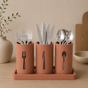 May include: A set of three cylindrical terracotta-colored utensil holders arranged on a matching rectangular tray. Each holder features a cutout silhouette of the specific utensil it contains: forks in the left, knives in the center, and spoons in the right.
