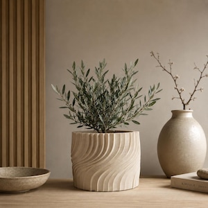 May include: A cylindrical beige planter with a vertical wavy textured pattern containing a small green olive tree. The planter sits alongside a speckled beige bowl, a tall textured beige vase with a thin branch, and a book titled KINFOLK JAPAN.