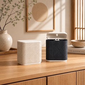 May include: Two rectangular waste bins with a textured geometric maze pattern on the exterior. One bin is cream-colored and closed, while the other is dark charcoal gray with an open white lid, revealing a small internal hinge mechanism.