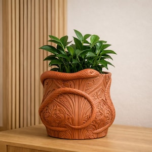 May include: A terracotta-colored cylindrical planter featuring a relief sculpture of a snake coiled around the exterior. The surface is decorated with intricate embossed floral and leaf patterns. A vibrant green leafy plant grows from the top.