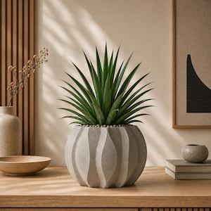 May include: A green spiky plant in a gray, textured, round pot with a wavy design. The pot sits on a wooden surface. The plant is surrounded by small, round pebbles. The background includes a wooden wall panel and a framed abstract art piece.