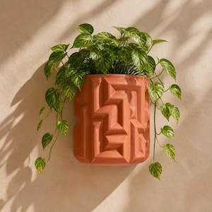 May include: A terracotta-colored wall-mounted planter featuring a geometric, maze-like relief pattern on its exterior surface. A trailing green and yellow variegated pothos plant grows from the top, with vines cascading down the sides.