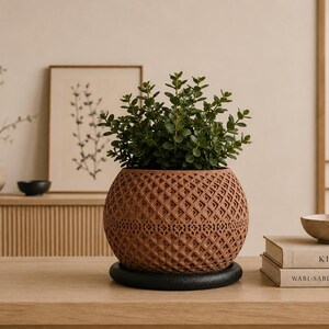 May include: A spherical terracotta-colored planter with a detailed geometric lattice pattern on its exterior. The planter sits on a circular black base and contains a lush green plant with small, rounded leaves, creating a textured appearance.
