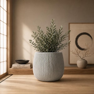 May include: A light gray, rounded planter with a vertical ribbed texture containing a lush green leafy plant. The planter has a wide, curved body and a matte finish, providing a neutral base for the dense foliage extending from the top opening.