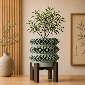 May include: A sage green cylindrical planter with a repeating geometric lattice pattern, featuring three distinct horizontal tiers. The planter is supported by a dark brown wooden stand with four legs and holds a small green leafy plant.