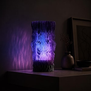 May include: A cylindrical lamp with a textured, pleated exterior that emits a vibrant purple and blue light. The structure features vertical folds creating a rippled effect, casting soft, patterned shadows on the surrounding area.
