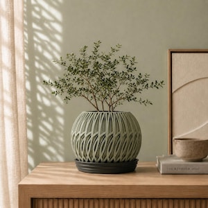 May include: A small green leafy plant potted in a spherical sage green planter with a decorative lattice-style cutout exterior. The planter sits on a dark circular saucer, showcasing a textured, woven-like pattern that wraps around the entire base.