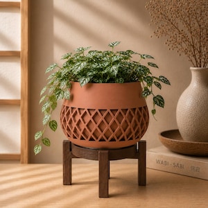 May include: A terracotta-colored planter with a lattice-patterned base, resting on a dark brown wooden four-legged stand. The planter holds a trailing green plant with variegated leaves, featuring small, heart-shaped foliage with silver patterns.