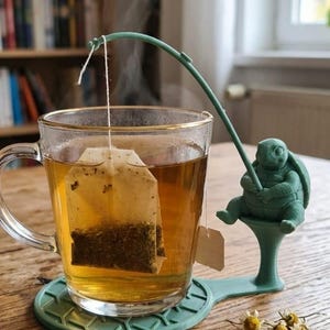 May include: A clear glass mug filled with amber tea and a submerged tea bag, resting on a sage green coaster. A sage green turtle figurine sits on an extension of the coaster, holding a fishing rod that suspends the tea bag string above the liquid.