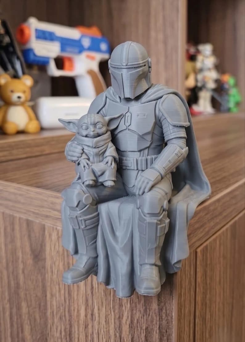 Puede incluir: Una figura gris impresa en 3D de un personaje blindado con casco sentado en un borde mientras sostiene a una peque&ntilde;a criatura de orejas grandes y ojos abiertos en su regazo. La figura lleva una capa y una armadura detallada en el pecho.