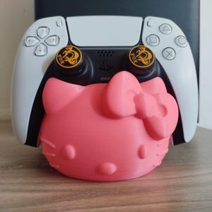 Puede incluir: Un soporte rosa con forma de Hello Kitty que sostiene un mando de juego blanco y negro. El mando tiene botones negros y detalles dorados. El soporte está sobre una superficie de madera.