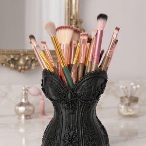 Peut inclure: Un porte-pinceaux de maquillage noir en forme de corset avec des détails en relief floraux et des volutes complexes. Le support est rempli de divers pinceaux de maquillage aux manches métalliques dorés, rose doré et roses avec des poils doux.