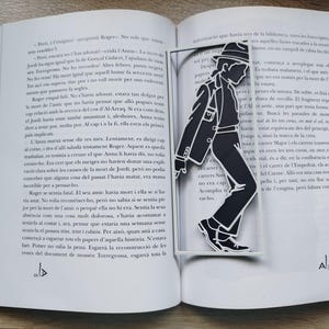 Puede incluir: Un marcapáginas de metal negro con la silueta recortada de una persona con sombrero fedora y traje, representada en una pose dinámica al caminar. El marcapáginas es rectangular con un marco fino y está colocado dentro de un libro abierto.