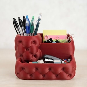 Può includere: Un organizer da scrivania rosso scuro con un design trapuntato a bottoni. L'organizer presenta una sezione cilindrica portapenne e uno scomparto rettangolare per cancelleria, contenente penne, matite, foglietti adesivi, clip e una cucitrice.
