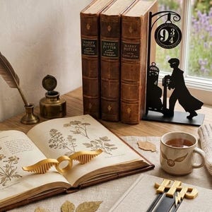 Può includere: Una scena a tema Harry Potter. Sono esposti tre libri rilegati in pelle, un anello con il Boccino d'Oro e un cartello della piattaforma 9 3/4. Una tazza con un design del Boccino d'Oro, una penna e un segnalibro creano un'atmosfera magica.