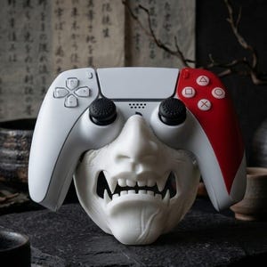 May include: A white and red video game controller with a white, detailed mask of a Japanese demon. The controller has black joysticks and a directional pad. The mask has sharp teeth and is placed below the controller.
