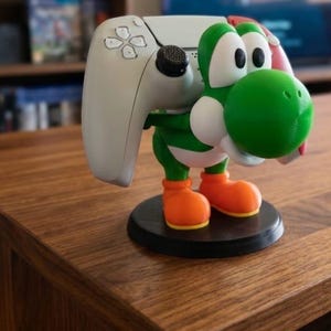 Könnte beinhalten: Eine grüne, weiße und orangefarbene Yoshi-Figur, die einen weißen Gamecontroller hält. Die Figur steht auf einem schwarzen, kreisförmigen Sockel, und der Controller ist so positioniert, als würde er gehalten. Die Figur steht auf einer Holzoberfläche.