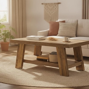 May include: A rectangular wooden coffee table with a natural brown finish, featuring four angled legs and a lower storage shelf. The tabletop holds two stacked books, a small dried plant stem, and a white ceramic mug on a wooden coaster.