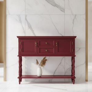 May include: A deep red console table featuring two central drawers with round brass knobs, two side cabinets with brass handles, and four turned legs. A lower shelf connects the legs, and a white vase with dried grass sits on the shelf.