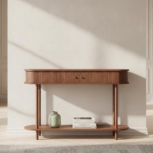 May include: A brown wooden console table featuring a rounded rectangular top with a slatted front design, two small circular knobs, and a lower shelf. The lower shelf holds a light green ceramic vase and a stack of three white books.
