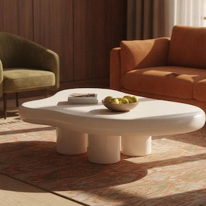 May include: A low-profile, off-white coffee table with an irregular, organic, cloud-like shape. The table features a smooth, rounded surface supported by three thick, cylindrical legs. A small stack of books and a bowl of green pears sit on top.