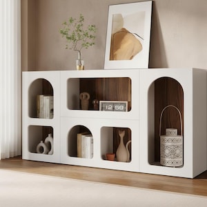 May include: A white sideboard featuring a combination of arched and rectangular open shelving units with a dark wood interior. The unit contains various decorative items including books, small figurines, a digital clock displaying 12:59, and a white lantern.