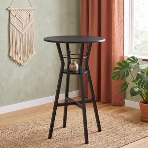 May include: A tall, round black bar table featuring a smooth tabletop and a lower circular shelf supported by four angled legs. The structure includes a metal ring accent between the tabletop and the shelf, providing a stable base for elevated seating.