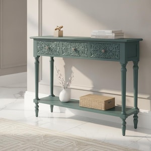 May include: A dark teal console table featuring three drawers with intricate carved floral patterns and small round knobs. The table has four slender turned legs and a lower shelf. On top are two stacked books and a gold object, with a white vase and a woven box on the shelf.