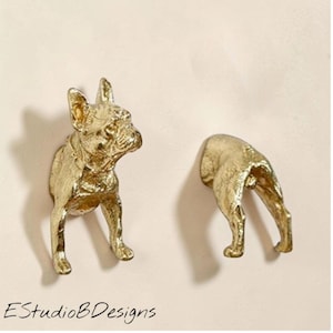 Pode incluir: Duas esculturas douradas de buldogue francês. Uma escultura retrata a cabeça e as patas dianteiras do cão, enquanto a outra mostra a metade traseira. As esculturas são montadas em uma superfície clara. O texto "EstudioBDesigns" está na parte inferior.