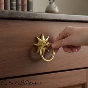 Puede incluir: Un tirador de cajón dorado en forma de estrella con un anillo circular fijado a un frente de cajón de madera. El herraje presenta una placa trasera de estrella de múltiples puntas y un anillo liso y redondeado. El texto EStudioBDesigns es visible.