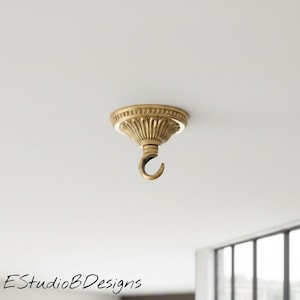 Könnte beinhalten: Ein Deckenhaken aus Messing mit einer kreisförmigen Basis, einem dekorativen geriffelten Design und einem Perlenrand. Das Beschlagteil hat ein warmes metallisches Finish und einen nach unten gebogenen Haken zur Montage an der Decke.