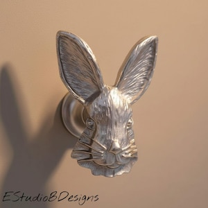 May include: A silver-toned metal cabinet knob shaped like a rabbit head with long, upright ears, detailed fur texture, and prominent whiskers. The hardware features a circular base mount and is positioned against a solid, neutral-colored surface.