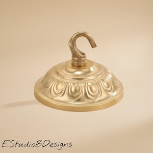 May include: A circular brass ceiling canopy featuring a central hook and a decorative embossed petal pattern around the dome. The hardware has a polished metallic finish and is designed for mounting light fixtures to a ceiling surface.