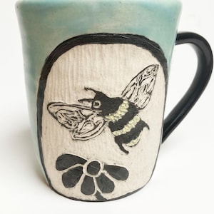 Peut inclure: Une tasse en céramique avec un vernis bleu clair et une anse noire. L'avant présente un panneau ovale texturé de couleur crème avec une illustration sculptée en noir et blanc d'un bourdon au-dessus d'une fleur, le tout dans un cadre noir.